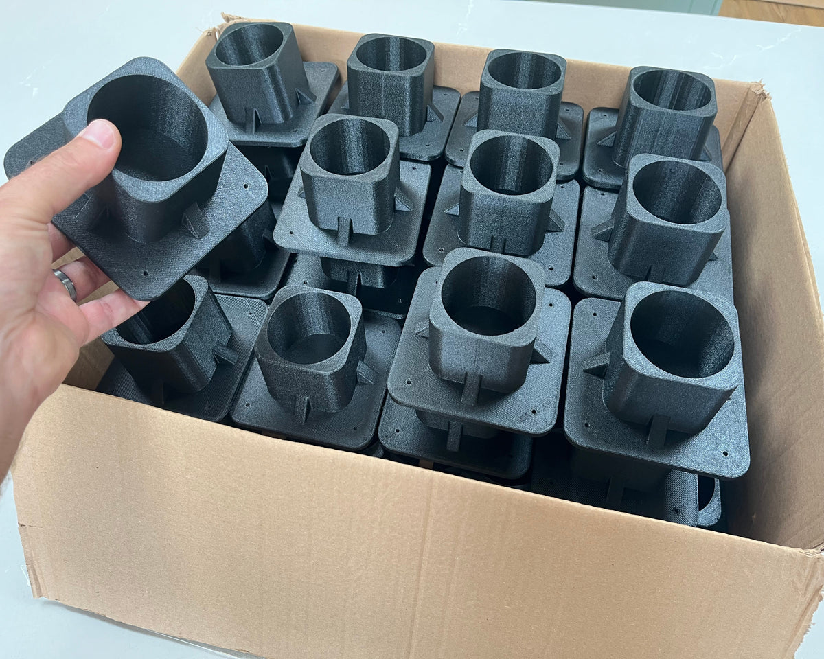 A hand holds a strong plastic mounting flange for tubing in front of a cardboard box filled with multiple identical components.