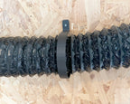 A black corrugated flexible duct hose with a matching black mounting bracket, laid horizontally on a light brown wood OSB board background.