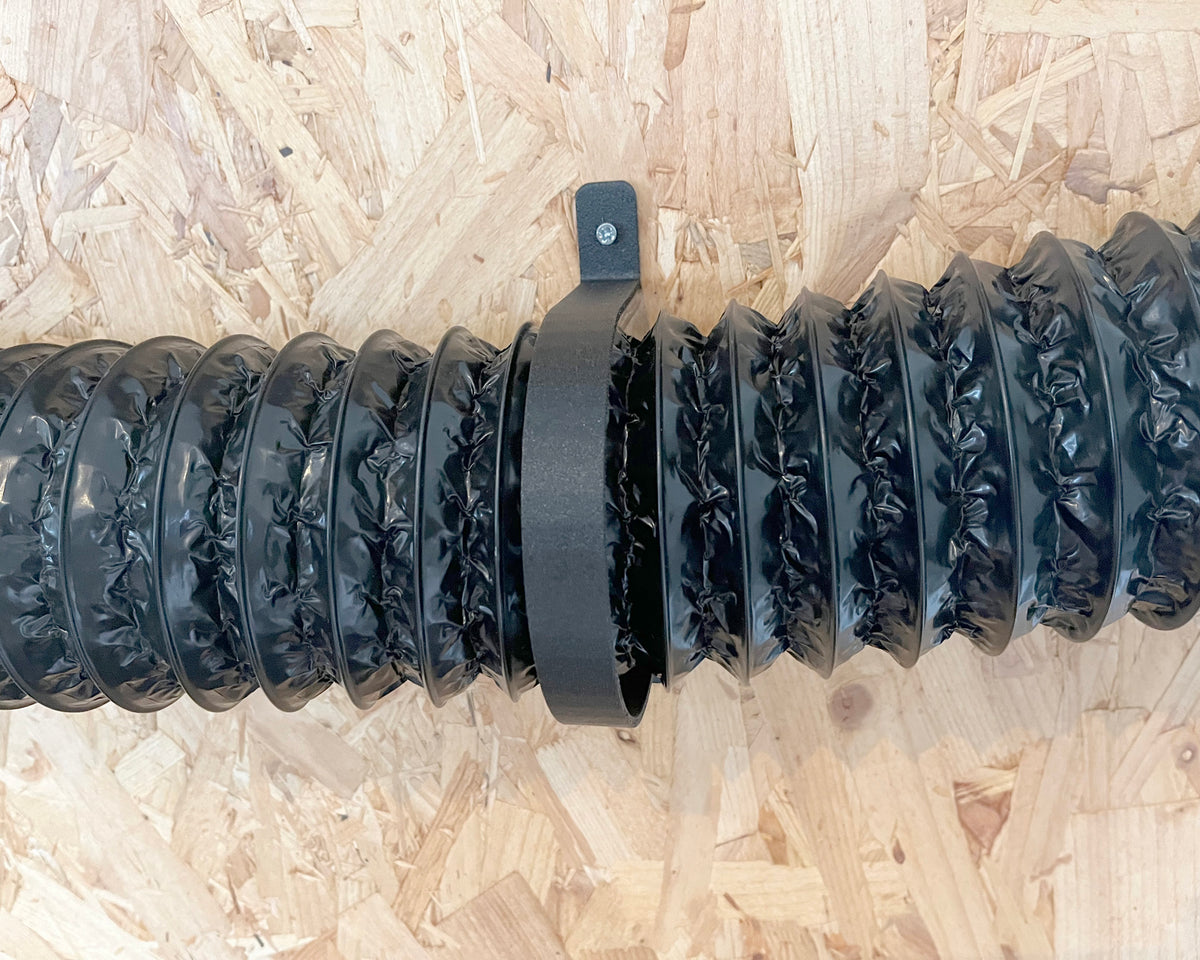 A black corrugated flexible duct hose with a matching black mounting bracket, laid horizontally on a light brown wood OSB board background.