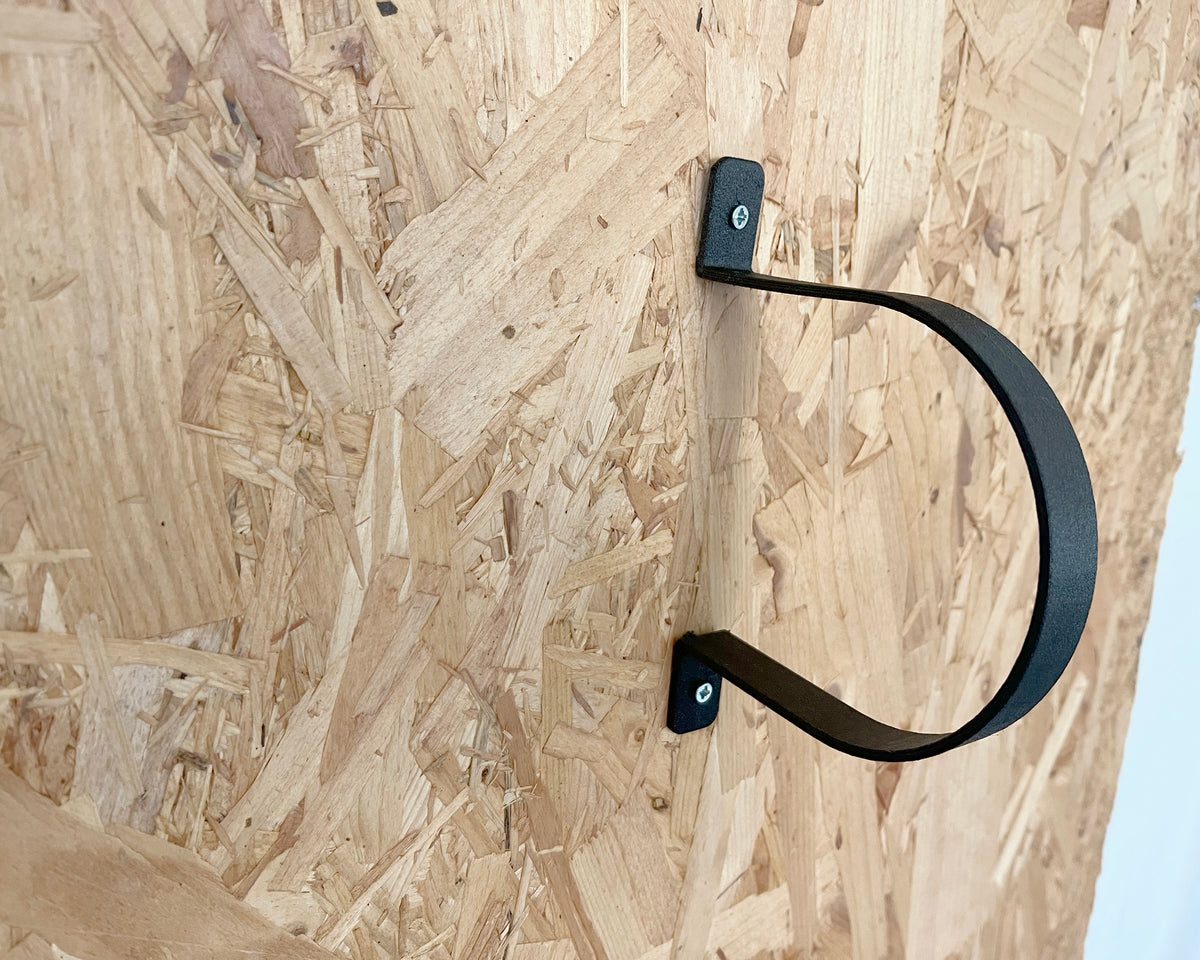 A black metal U-shaped wall hook bracket mounted on an OSB board wall.